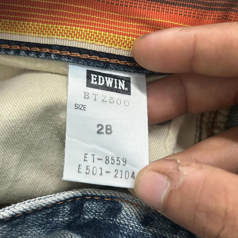 May include: Close-up of a white tag with the brand name "EDWIN" in black. The tag also displays the size "28" and other product codes. The tag is attached to a pair of blue denim jeans.