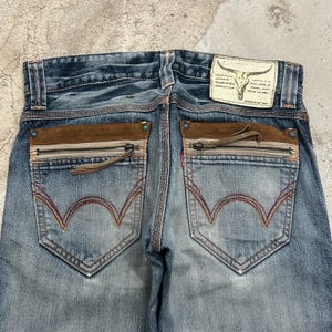 May include: Close-up of the back of blue denim jeans with brown suede accents on the pockets. The jeans have a zipper detail on the pockets and a leather patch with a bull skull logo.