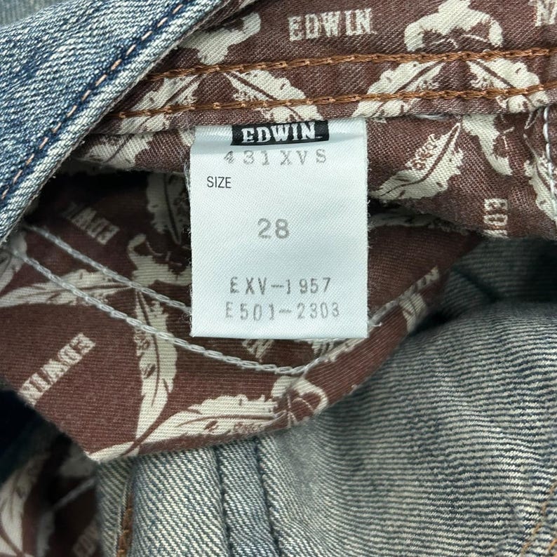 May include: Close-up of a denim jeans label with the brand name "EDWIN" visible. The label indicates a size of 28. The interior lining of the jeans features a brown and white patterned fabric. The jeans are a light blue wash.