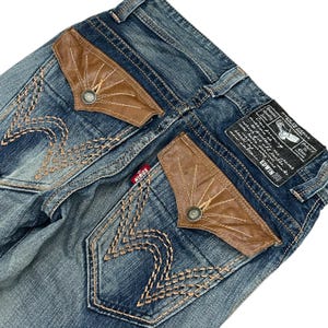 May include: Blue denim jeans with brown leather pocket detailing and decorative stitching. The back pockets have a unique design with a button closure. A small red brand tag is visible.