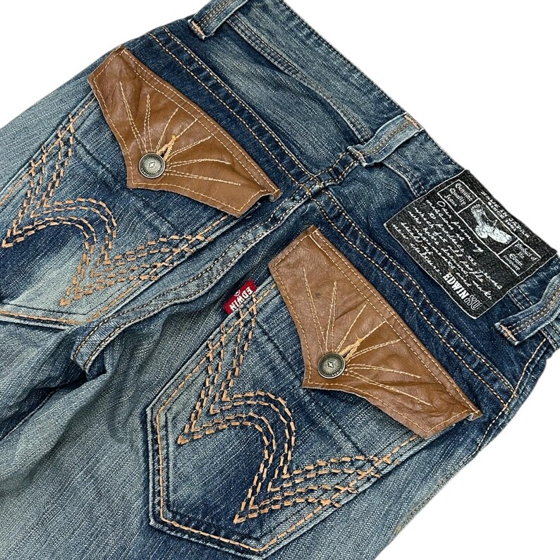 May include: Blue denim jeans with brown leather pocket detailing and decorative stitching. The back pockets have a unique design with a button closure. A small red brand tag is visible.