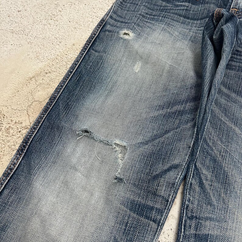 May include: Close-up of distressed blue denim jeans. The jeans have a faded wash with visible wear and tear, including a large rip on the knee area. The fabric shows signs of fading and distressing, giving it a vintage look. The jeans are a classic five-pocket style.
