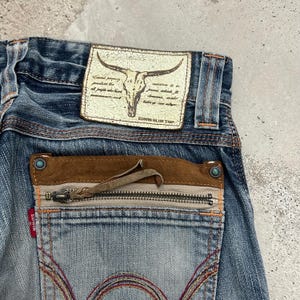 May include: Close-up of blue denim jeans with a distressed look. Features a leather patch with a bull skull graphic and text. The back pocket has a zipper and brown suede detailing. The brand name "EDWIN BLUE TRIP" is visible.