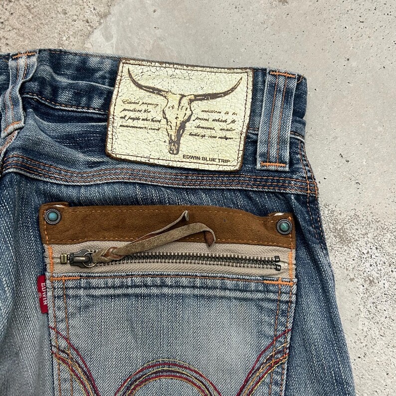 May include: Close-up of blue denim jeans with a distressed look. Features a leather patch with a bull skull graphic and text. The back pocket has a zipper and brown suede detailing. The brand name "EDWIN BLUE TRIP" is visible.