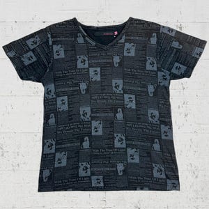 以下が含まれることがあります： 新聞プリントのデザインが施された黒いVネックTシャツ。 "We Dream The Future" や "With The Tone Of Light" などのフレーズを含むテキストと画像が含まれています。 シャツは柔らかい素材で作られています。