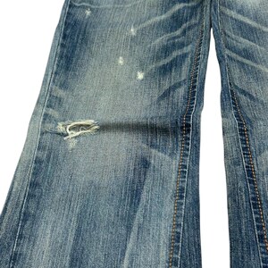 May include: Close-up of light blue denim jeans with distressed details. The jeans have a ripped section on the left leg and visible stitching. The denim has a faded appearance with lighter patches.