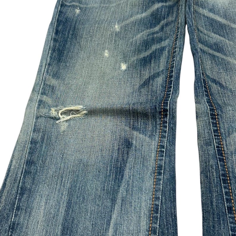 May include: Close-up of light blue denim jeans with distressed details. The jeans have a ripped section on the left leg and visible stitching. The denim has a faded appearance with lighter patches.