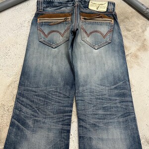May include: Blue denim jeans with a faded wash and distressed details. Features include brown suede-like accents on the back pockets, zipper details, and a leather patch with a bull skull logo. The jeans have a classic five-pocket design.