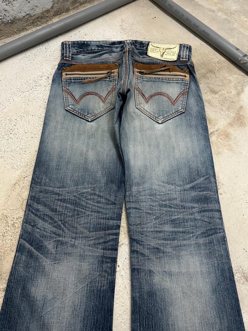 May include: Blue denim jeans with a faded wash and distressed details. Features include brown suede-like accents on the back pockets, zipper details, and a leather patch with a bull skull logo. The jeans have a classic five-pocket design.