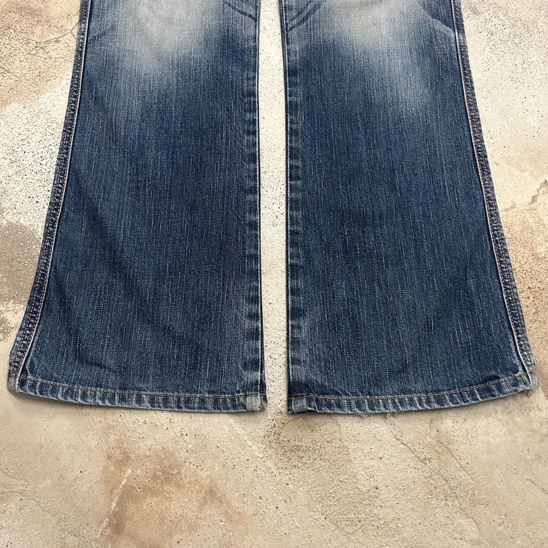 May include: A pair of blue denim jeans with a flared leg design. The jeans have a faded wash effect and visible stitching along the seams. The bottom hem is finished with a classic rolled edge.