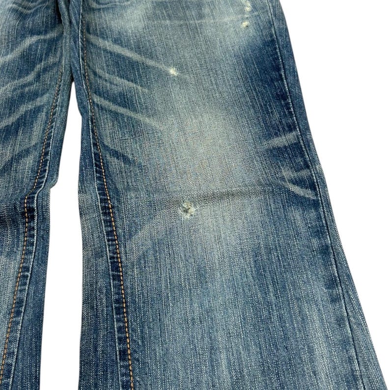 May include: Close-up of blue denim jeans. The jeans have a faded wash with distressed details, including a small hole and worn areas. Stitching is visible along the seams.