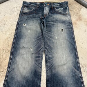 May include: Faded blue denim jeans with distressed details, including rips and worn patches. The jeans feature orange stitching, classic five-pocket styling, and a button and zipper closure. The denim has a washed appearance.