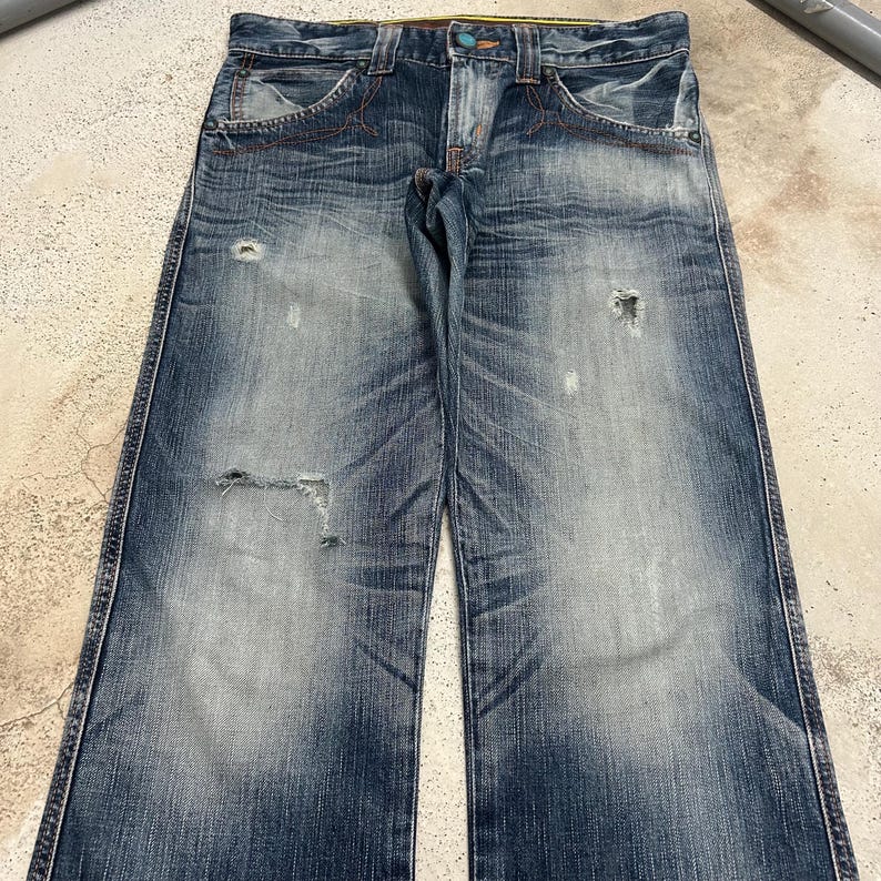 May include: Faded blue denim jeans with distressed details, including rips and worn patches. The jeans feature orange stitching, classic five-pocket styling, and a button and zipper closure. The denim has a washed appearance.