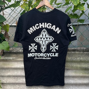 May include: Black t-shirt with white graphic text and design. The text reads "MICHIGAN MOTORCYCLE Custom Builder" with a cross and skull design. The shirt is hanging on a wooden hanger.