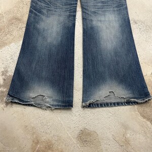 May include: Faded blue denim jeans with a flared leg design. The jeans show wear and tear, with distressed areas near the hem. The denim has a washed appearance, with lighter shades in the center of the legs.