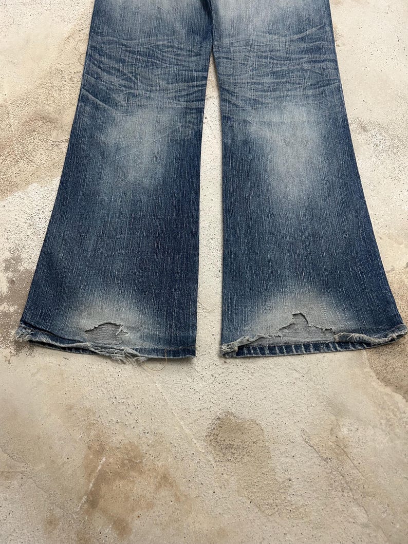 May include: Faded blue denim jeans with a flared leg design. The jeans show wear and tear, with distressed areas near the hem. The denim has a washed appearance, with lighter shades in the center of the legs.