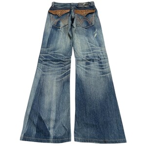 May include: Blue denim jeans with a wide leg design. The jeans feature brown leather-like accents on the back pockets and a distressed wash. The back pockets have a decorative stitch pattern.