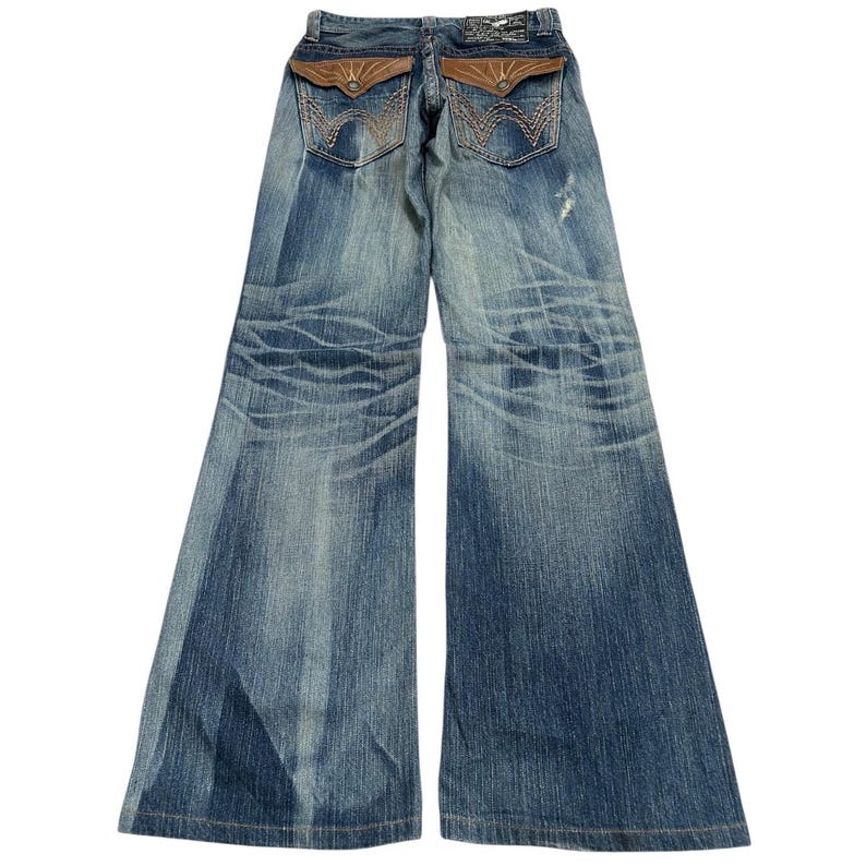 May include: Blue denim jeans with a wide leg design. The jeans feature brown leather-like accents on the back pockets and a distressed wash. The back pockets have a decorative stitch pattern.