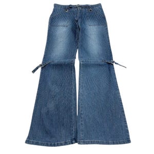 May include: Blue denim trousers featuring a vertical pinstripe pattern, a wide-leg silhouette, and two front pockets with silver-toned metal rings. The trousers include adjustable side straps with metal buckles at the mid-thigh and a waist belt loop.