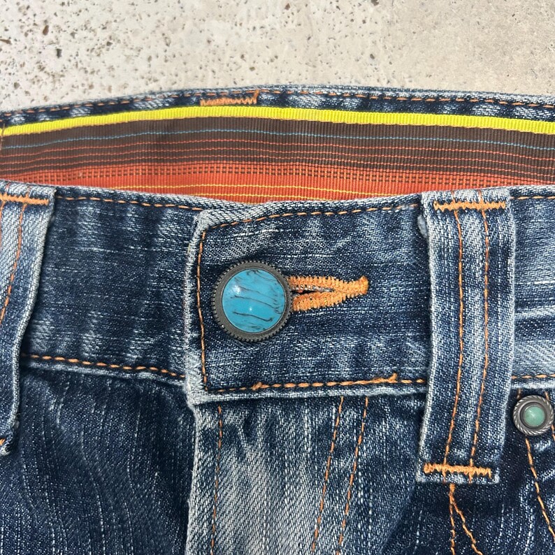 May include: Close-up of blue denim jeans with orange stitching. The jeans feature a turquoise button and a decorative waistband with yellow, orange, and brown stripes. The button is surrounded by a dark metal ring.