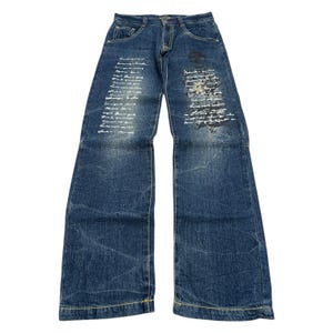 May include: Blue denim jeans with a wide leg design. The jeans feature white script and a skull graphic on the front. The jeans have a classic five-pocket design and yellow stitching.