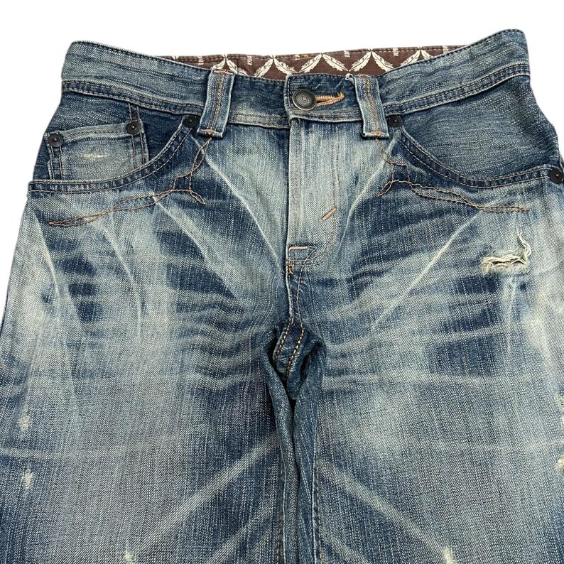 May include: Close-up of faded blue denim jeans with distressed details. The jeans feature a button closure, belt loops, and pockets. The fabric has a worn, textured appearance with light and dark blue washes, and a brown patterned waistband.