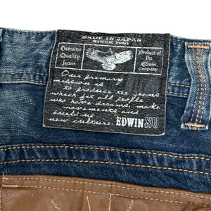 May include: Close-up of blue denim jeans with a leather patch. The patch features text that reads "MADE IN JAPAN SINCE 1961" and "Genuine Quality Jeans." An eagle graphic is also visible. The brand name "EDWIN XU" is displayed.