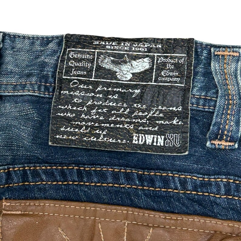 May include: Close-up of blue denim jeans with a leather patch. The patch features text that reads "MADE IN JAPAN SINCE 1961" and "Genuine Quality Jeans." An eagle graphic is also visible. The brand name "EDWIN XU" is displayed.