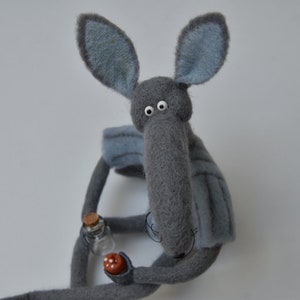 Puede incluir: Un animal de peluche gris de fieltro con orejas grandes y un hocico largo. El juguete tiene grandes ojos blancos y sostiene una pequeña botella de vidrio en su mano.