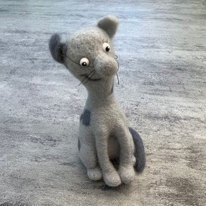 Puede incluir: Figura de gato de fieltro gris hecha a mano con un diseño caprichoso. El gato tiene ojos grandes y expresivos, un cuello largo y una cola pequeña. Presenta una oreja gris oscuro y una mancha gris oscuro en la espalda. El gato está sentado sobre una superficie gris.