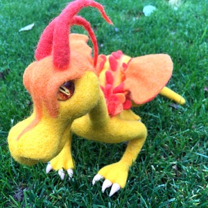 Puede incluir: Un dragón de fieltro amarillo y naranja con cuernos rojos y un ojo blanco. El dragón está posado sobre sus patas delanteras con sus alas extendidas detrás de él. El dragón está sentado en un lecho de hierba verde.