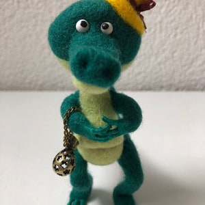 Puede incluir: Un cocodrilo de fieltro verde con grandes ojos blancos y un sombrero de fieltro amarillo con un lazo rojo. El cocodrilo sostiene una pequeña cadena dorada adornada con una bola decorativa en el extremo.