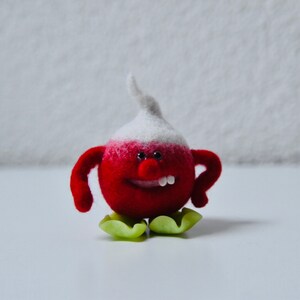 Puede incluir: Un juguete de fieltro rojo y blanco con una cara sonriente y pies verdes. El juguete tiene forma de rábano.