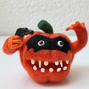 Puede incluir: Una calabaza de fieltro con una máscara negra para los ojos, dientes blancos y una pistola de juguete negra. La calabaza es naranja y tiene un tallo verde.