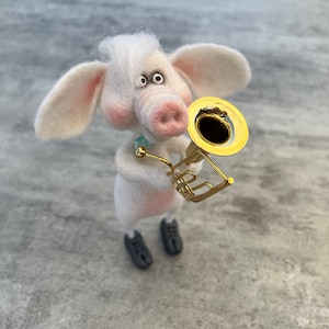 Puede incluir: Un cerdo de fieltro blanco con zapatos negros toca una trompeta dorada. El cerdo tiene una nariz rosada y ojos grandes y oscuros.