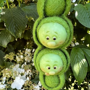 Puede incluir: Tres vainas de guisantes de fieltro verde con caras lindas. Las vainas de guisantes están colgando de una rama verde frondosa.