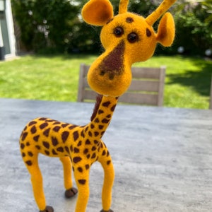Puede incluir: Un juguete de jirafa de fieltro amarillo y marrón con ojos grandes y cuello largo. La jirafa está de pie sobre una superficie gris.