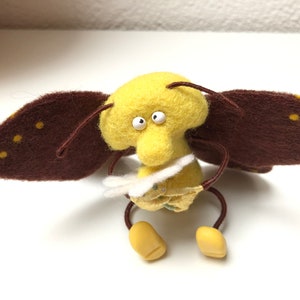 Puede incluir: Una mariposa de fieltro amarilla con alas marrones y manchas amarillas. La mariposa tiene ojos grandes y blancos y sostiene una flor de fieltro blanca.