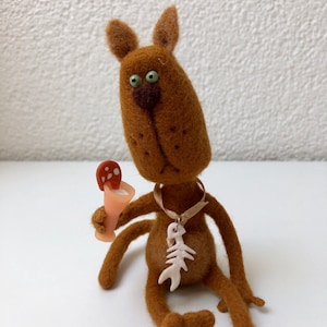 Puede incluir: Un juguete de gato de fieltro marrón con ojos verdes, nariz marrón y collar de hueso blanco. El gato sostiene un pequeño hongo rojo y blanco en su pata.