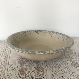 Puede incluir: Un tazón grande de cerámica de color crema con un esmalte moteado azul y blanco. El tazón tiene un borde ancho y redondeado y una superficie lisa ligeramente texturizada.