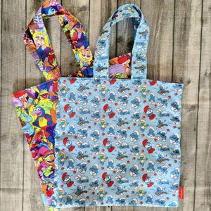 Könnte beinhalten: Zwei Stofftragetaschen mit Henkeln. Eine Tasche hat einen hellblauen Hintergrund mit einem sich wiederholenden Muster aus kleinen blauen Schlümpfen. Die zweite Tasche liegt darunter und zeigt eine bunte Collage verschiedener Muppet-Figuren.
