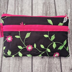 Puede incluir: Bolsa negra bordada con cremallera rosa y diseño floral. La bolsa presenta una cremallera rosa y un tirador en forma de hoja. El bordado floral incluye flores rosas y hojas verdes.