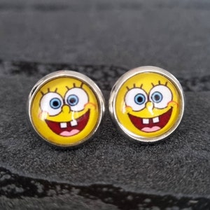 Peut inclure: Une paire de boucles d'oreilles en argent avec un visage de dessin animé jaune aux yeux bleus et un grand sourire. Le visage est celui d'un personnage de dessin animé populaire.