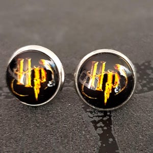 Puede incluir: Un par de pendientes de botón redondos con un engaste de metal plateado y un cabujón de cristal negro. El centro de cada pendiente muestra las letras HP en amarillo dorado con una fuente estilizada de llamas sobre fondo negro.