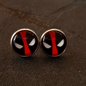 Może przedstawiać: Para srebrnych kolczyków z wzorem czerwono-czarnej maski Deadpoola.