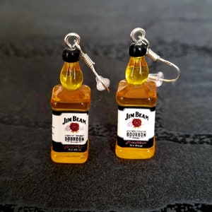 Könnte beinhalten: Ein Paar Miniatur-Ohrringe in Form von Jim Beam Bourbon-Flaschen. Die Ohrringe zeigen klare, bernsteinfarbene Flaschen mit schwarzen Verschlüssen und weißen Etiketten. Die Ohrringe haben silberfarbene Ohrhaken.