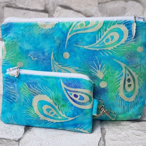Pode incluir: Duas bolsas com zíper azuis e verdes com um padrão paisley. A bolsa maior tem um amuleto de pena de prata preso ao puxador do zíper. A bolsa menor tem um amuleto de pena de prata preso ao puxador do zíper e um chaveiro de prata.