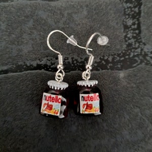 Könnte beinhalten: Ein Paar silberne Ohrhänger mit Miniatur-Gläsern mit einer braunen Flüssigkeit und einem weißen Etikett mit der Aufschrift "Nutella".