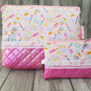 Könnte beinhalten: Zwei rosa Kosmetiktaschen. Die größere Tasche hat eine rechteckige Form, und die kleinere Tasche ist rechteckig. Beide Taschen haben einen gesteppten, irisierenden rosa Boden und eine weiße Spitzentrimmung. Der obere Teil der Taschen zeigt ein Muster aus Süßigkeiten.