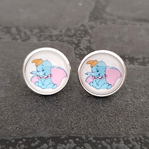 Może przedstawiać: Para srebrnych kolczyków z białym tłem i kreskówkowym obrazkiem Dumbo słonia z różowymi uszami i żółtym kapeluszem.